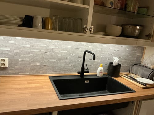 Kundens lysegrå selvklebende veggfliser montert som en bakvegg på et kjøkken over en svart vask.