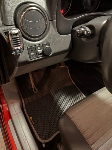 Recenzent pokazuje czarny dywanik samochodowy z jasnym obszyciem, dopasowany do miejsca kierowcy.