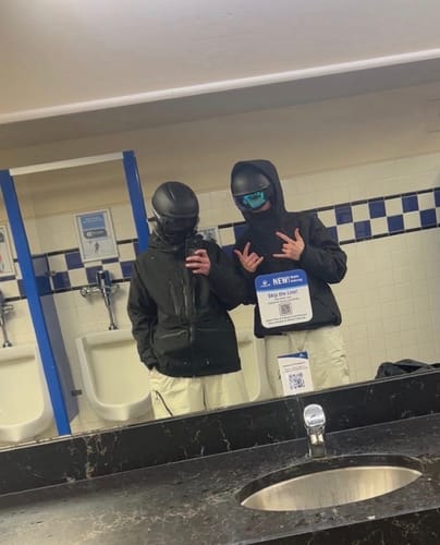 Reviewer takes a mirror selfie with another person in a restroom, both wearing black insulated snow jackets and helmets.