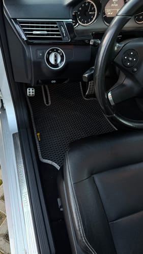 Recenzentka pokazuje czarny dywanik samochodowy przy fotelu kierowcy, z dopasowanym szarym obszyciem.