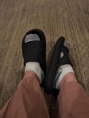 Reviewer's point of view showing their feet in black Cloudies Orthopedic Slippers, worn with socks indoors.
