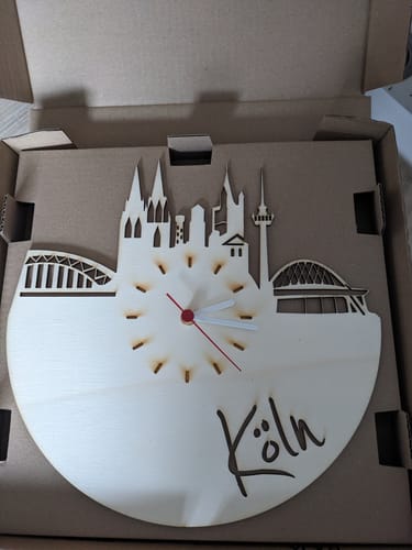 Kunde zeigt die Wanduhr aus Holz mit Kölner Skyline in der geöffneten Produktverpackung.