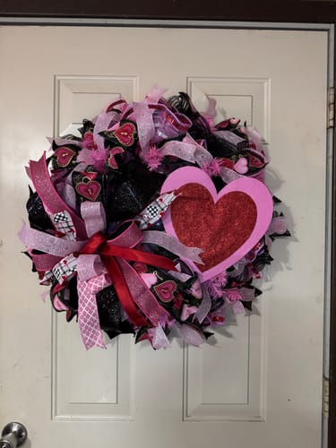 A customer's handmade Valentine's Day wreath hanging on a white door, featuring a prominent bow made with the red laser glitter ribbon.