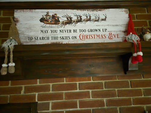 Reviewer's rustic Christmas sign displayed on a wooden mantelpiece, set above a brick fireplace and next to two gnomes.