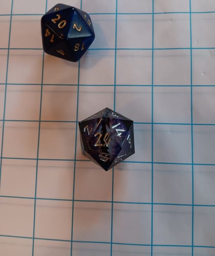 Customer's black and silver Enchanted Dragon D20 on a gridded mat, shown next to a standard blue die for scale.