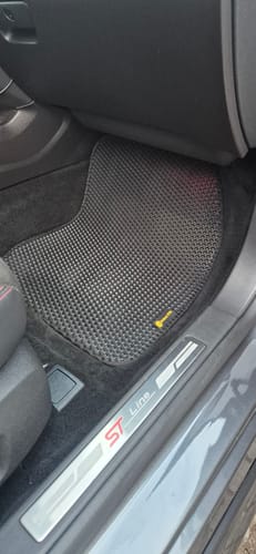 Recenzent pokazuje czarny, dopasowany dywanik samochodowy po stronie kierowcy w swoim aucie.