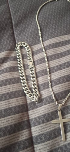 El cliente muestra su Pulsera Cuban 8mm plateada junto a un collar con una cruz sobre una tela a rayas.