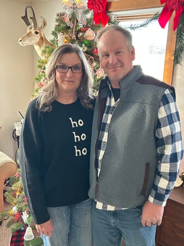 Customer wearing the black Classic Ho Ho Ho Sweater while standing next to another person by a Christmas tree.