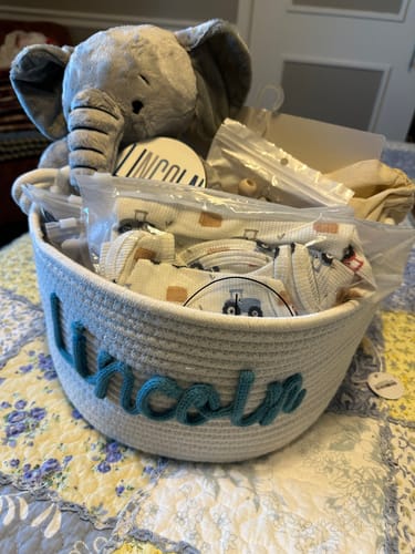 Customer's white woven basket personalized with the name 'Lincoln' in teal script, filled with baby gifts on a quilt.