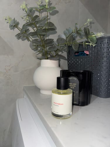 Customer's bottle of Moonbeam perfume sitting on a white countertop next to a vase of greenery and a candle.
