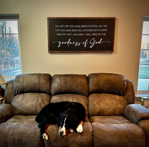 Customer's black Goodness Of God Faith Sign hung in a living room over a couch where a dog is sleeping.
