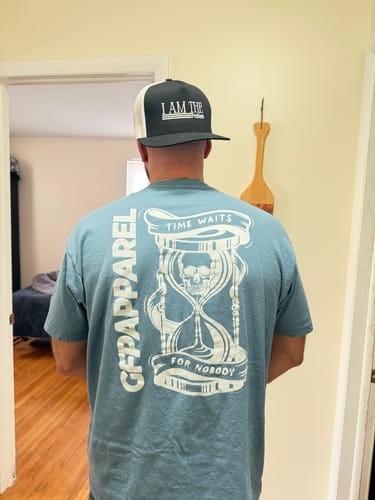 Reviewer wearing the back of the blue TIME TEE, showing the large "Time Waits For Nobody" hourglass graphic.