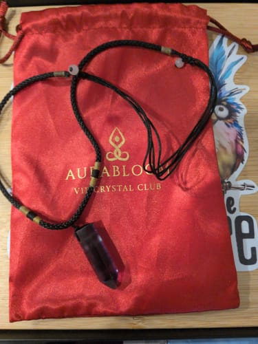 Reviewer's Natural Fluorite Necklace with a dark purple crystal pendant on a black cord, laid over its red satin pouch.