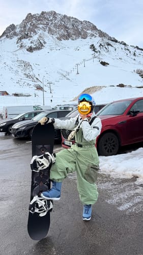 Reviewer posing in mint green baggy snow bibs with a snowboard in front of a snowy mountain.