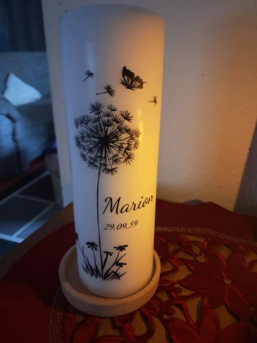 Rezensent zeigt die weiße Trauerkerze 'Pusteblume Schmetterling', personalisiert mit Namen und Datum, auf einem Tisch.