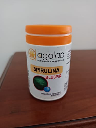 Recensore mostra il flacone di Spirulina BluSpir con tappo arancione, posato su un mobile di legno.