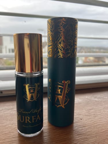 Customer's Surfari travel perfume bottle with a gold cap, pictured next to its dark green and gold case on a wooden surface.