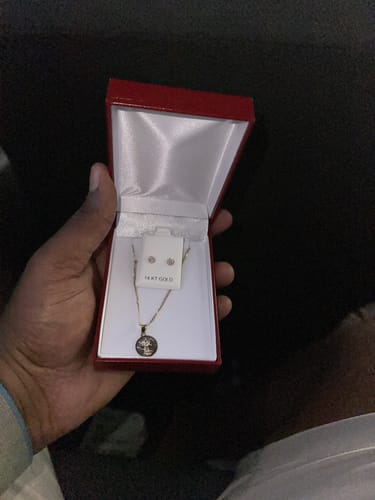 Customer's hand holds a red jewelry box open, revealing the Divino Niño pendant and earring set.