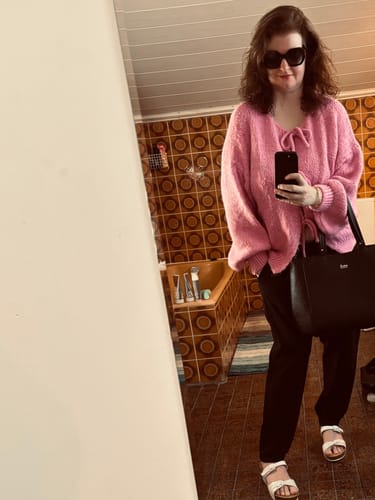 Rezensentin trägt den pinken Oversized Cardigan mit Schleifen, kombiniert mit einer schwarzen Hose und einer großen Tasche.