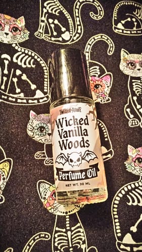 Reviewer's Wicked Vanilla Woods Perfume Oil rollerball on a dark fabric with decorative cat skeleton graphics.