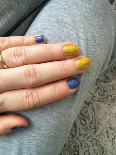 Hand einer Kundin, die die blauen 'Cold Night Cuddles' Nagelfolien mit goldenen Glitzernägeln kombiniert.