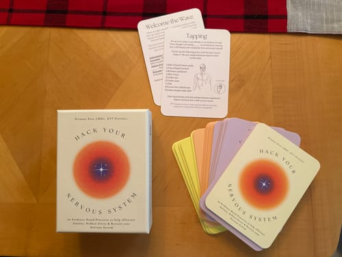 Reviewer's Hack Your Nervous System card deck with its box and several colorful cards fanned out on a wooden table.