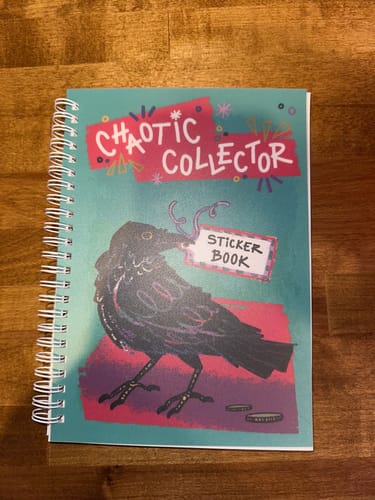 Reviewer's Chaotic Collector Reusable Sticker Book on a wooden table, showing the illustrated crow on its fun, teal cover.