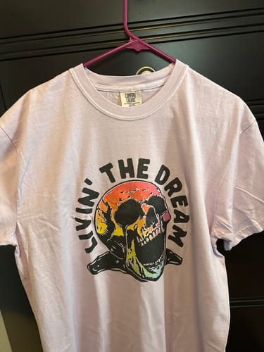 Customer's orchid Livin' The Dream tee with a multi-color skull graphic hanging on a hanger.