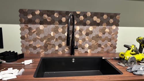 Anmelder som installerer Mocca hexagon selvklebende veggfliser over en svart kjøkkenvask.