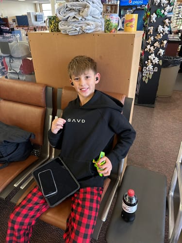Reviewer's child wearing the black Youth Oversized Hoodie with the front logo while sitting in an airport terminal.