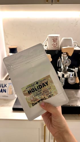 Reviewer holding a white pouch of the Holiday Blend coffee in front of an espresso machine.