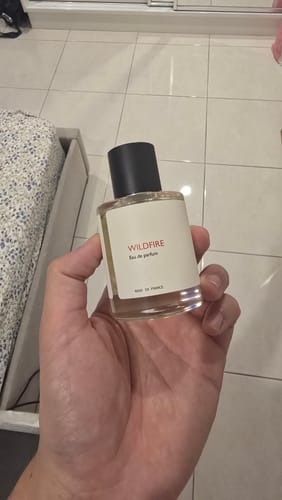 Reviewer holding a clear glass bottle of Wildfire eau de parfum, showing its white label and black cap.