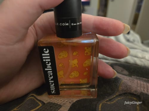 A reviewer holds the square bottle of Cheese Hangover perfume, showing the orange label with a whimsical cheese pattern.