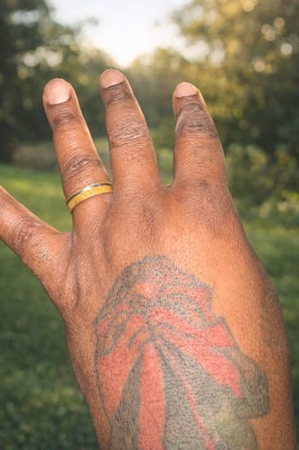 Customer's tattooed hand wearing The Oakhead ring with its tan wood inlay outdoors.