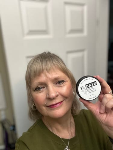 A smiling reviewer holds up the round, black tin of F-Balm.