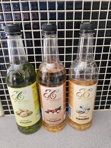 Reviewer's Macadamia Coffee Syrup bottle placed between Pistachio and Vanilla syrup bottles on a counter.