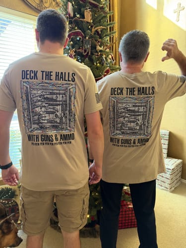 Two reviewers showing the back of their tan "Deck The Halls With Guns & Ammo" t-shirts by a Christmas tree.