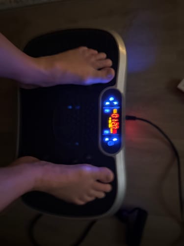 Anmelder står barfodet på sin Vibrationstræner, hvor det oplyste digitale display viser, at den er tændt.