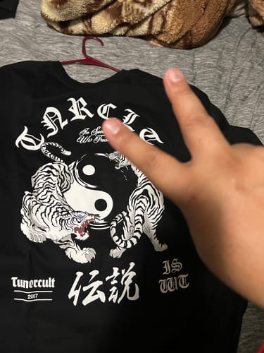 Reviewer showing the back of the black YIN-YANG TEE with its tiger graphic, while making a peace sign with their hand.
