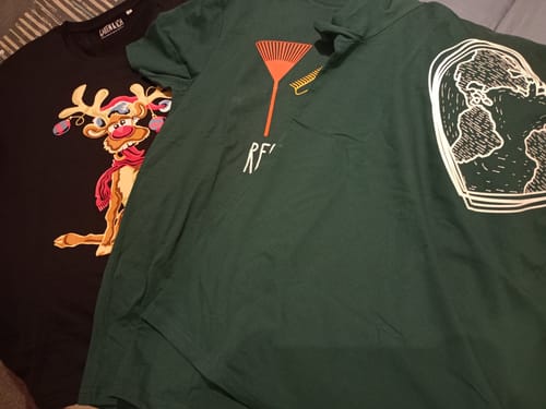 Kunde zeigt zwei erhaltene Shirts aus der Wundertüte: ein schwarzes Weihnachts-Shirt und ein dunkelgrünes T-Shirt mit Grafiken.