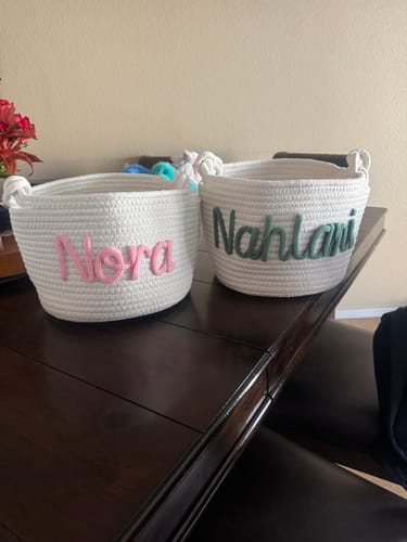 Reviewer's two personalized white woven baskets sitting on a wooden table, with the names 'Nora' and 'Nahlani' on them.