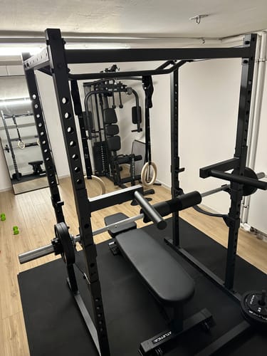 Rezensent zeigt das schwarze R5 Base Power Rack in einem Raum mit niedriger Decke, mit einer Hantelbank und Turnringen.