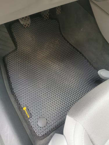 Recenzent pokazuje dopasowany dywanik samochodowy o strukturze plastra miodu, zamontowany po stronie kierowcy.