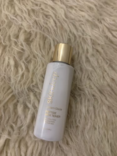 Customer's DREAM GLAZE Peptide Milk Toner bottle with a gold cap, resting on a fluffy cream-colored surface.