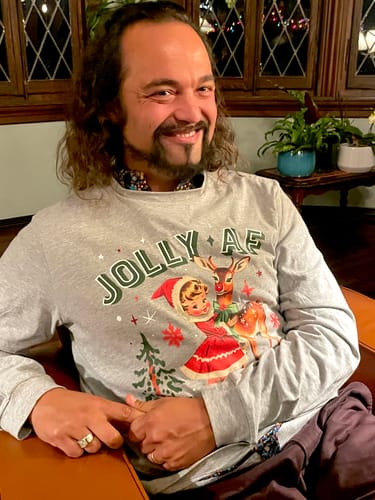 Reviewer smiling while sitting indoors and wearing the grey Jolly AF Long Sleeve Tee with a vintage Christmas graphic.