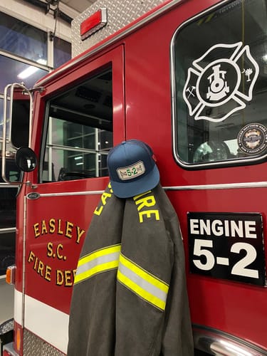 Customer's blue FLAT BILL HOSE HAT hanging on a firefighter's jacket in front of a red fire truck.
