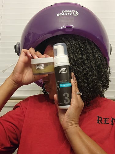 Reviewer holding the Tropical Oasis Hair Gel while sitting under a hooded dryer to set their curly hair.