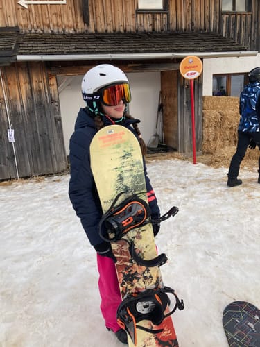 Reviewer in rose baggy snow pants standing in the snow while holding a snowboard.