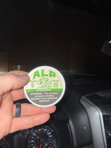 Customer holding a round can of ALP Spearmint Nicotine Pouches inside a car, in front of the dashboard.