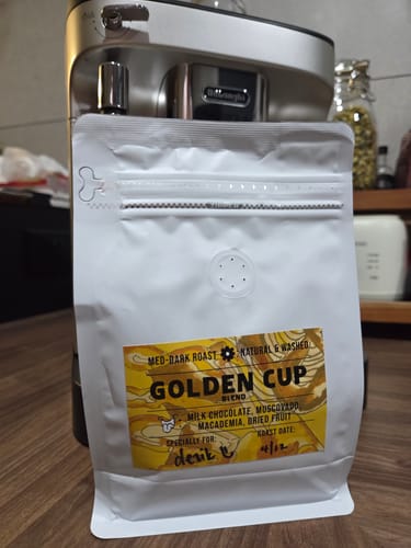 Reviewer's white bag of Golden Cup Blend coffee sitting on a counter in front of a coffee machine.